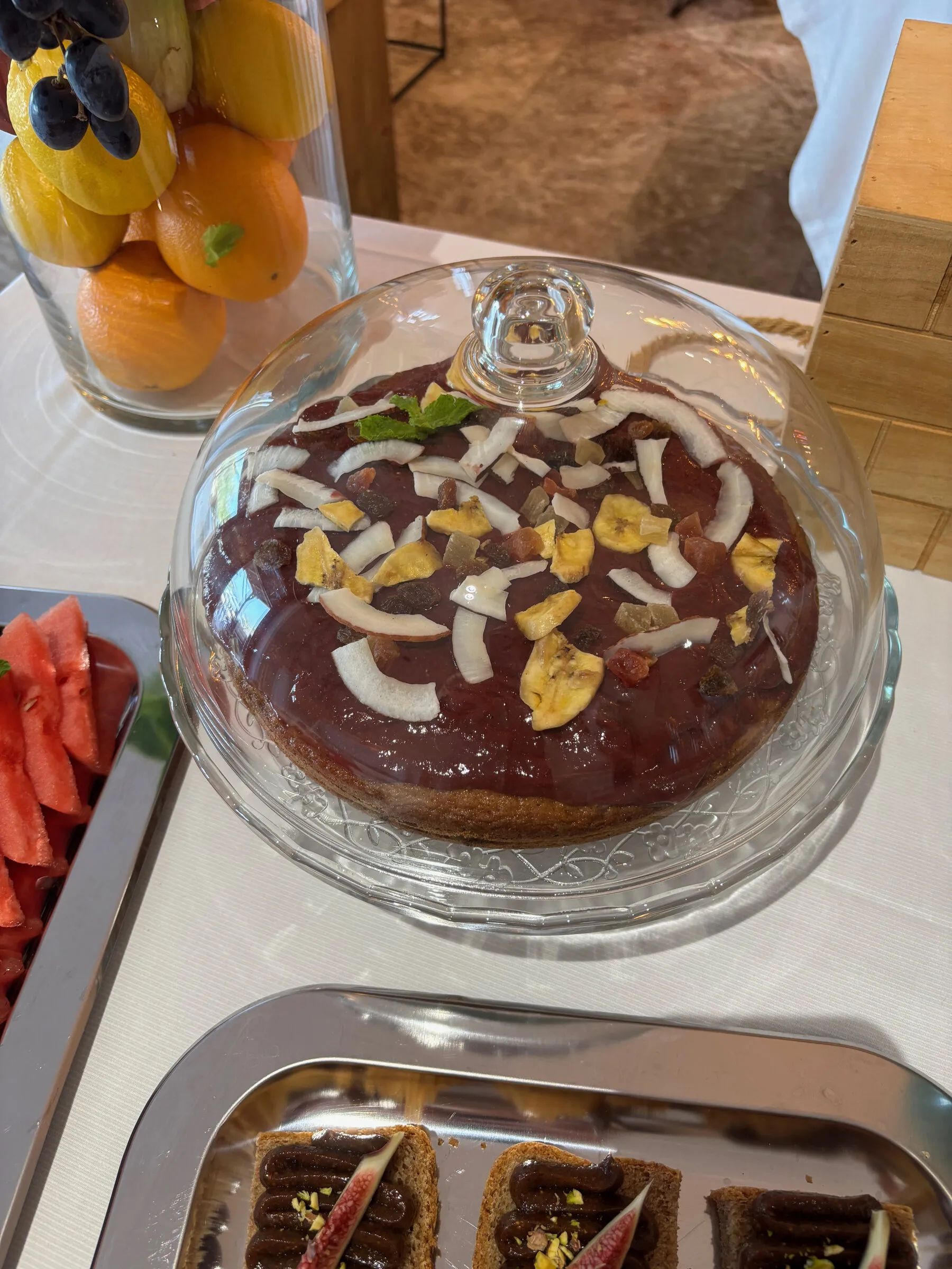 A cacao-glazed cake under a glass cloche, topped with coconut and dried fruit