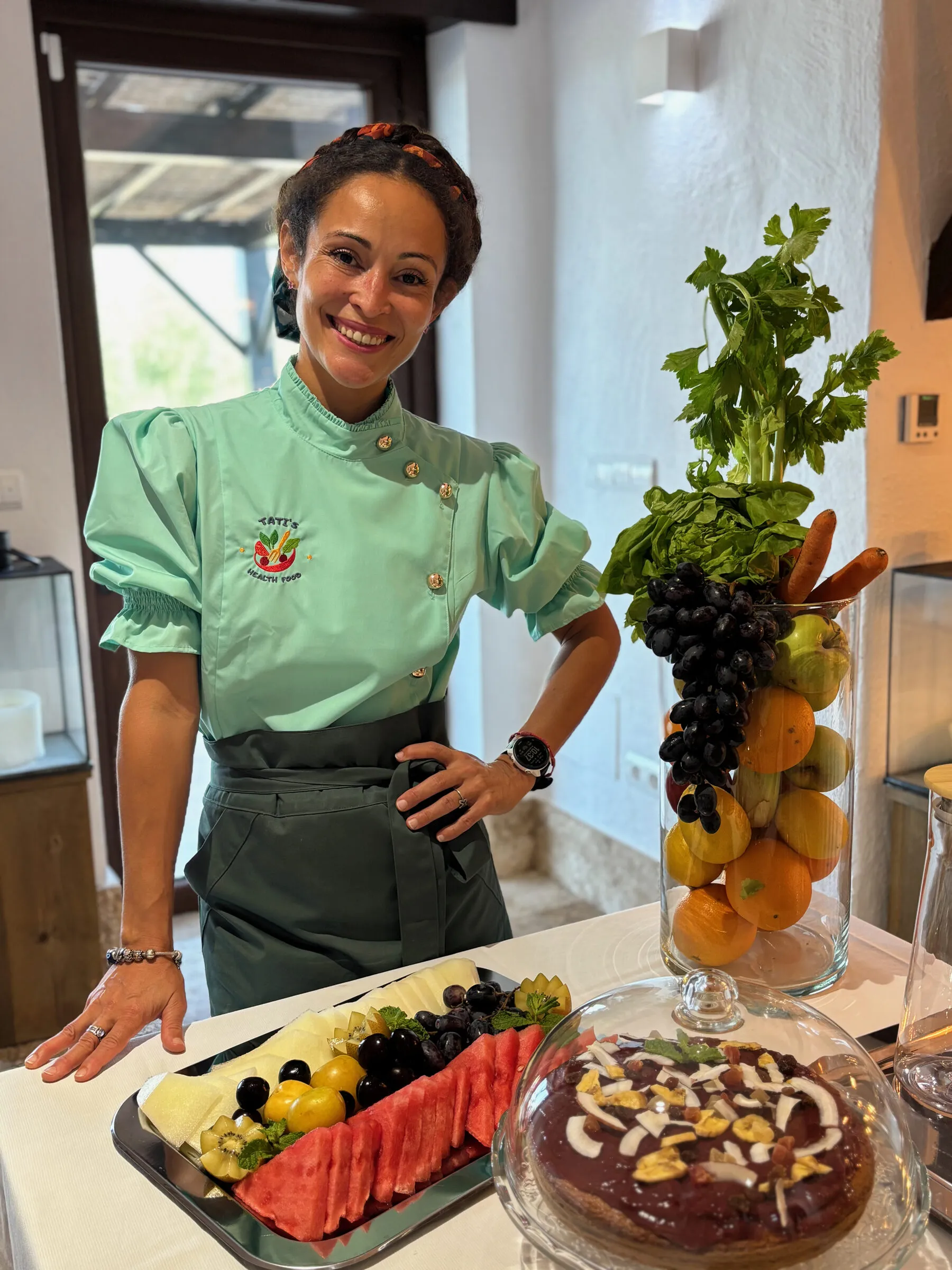 Tati in her chef whites beside a table of fruit, cake and fresh dishes
